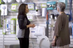 a pharmacy or medical supply store employee guiding an older female customer to view home safety products, NOVA HME products visible on back wall; The Bone Store