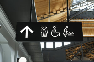an overhead sign inside an airport, depicting icons for family-friendly, disabled-friendly, and baby changing room restrooms with an arrow straight ahead