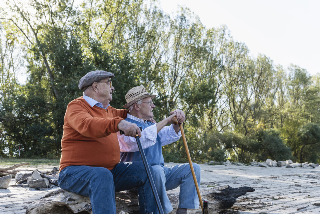 two older men who are friends sitting along the side of a waterway, each with his own type of cane for walking and balance, trees in the background