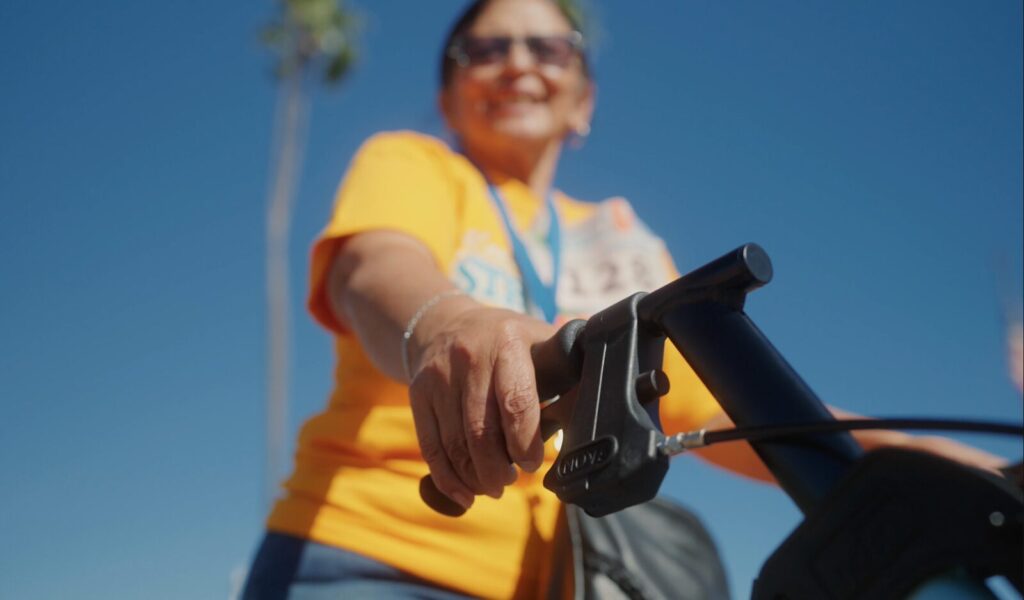 a woman smiles as walks with her NOVA rollator, the brake handle in focus, blue sky and palm trees in the background