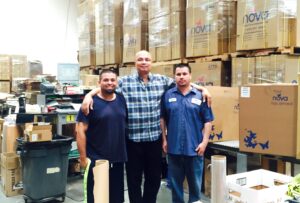 three men pose for a team photo in the NOVA Medical Products distribution warehouse; big brown NOVA boxes are towering in the background