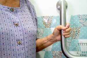 a mature woman holds onto a metal bathroom safety railing with one hand in a tiled bathroom setting
