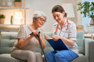 an older adult female sits, with walking cane in hand, on a home living room sofa with a female home nursing care worker reviewing medical paperwork on a clipboard