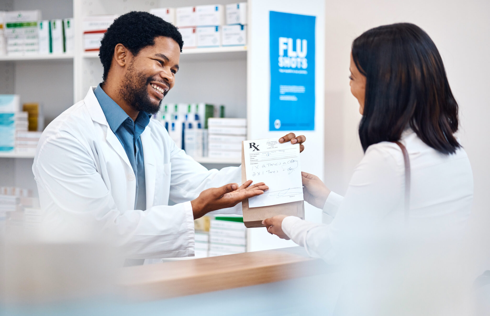 a male pharmacist behind the counter smiles as he hands a prescription medication bag to a female patient