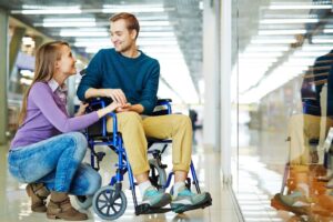 a young couple is smiling and holding hands in a shopping mall hallway; a woman squats down to hold hands with her male counterpart who is seated in a blue transport wheelchair