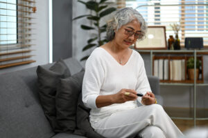 an older adult woman sits on a living room sofa checking her blood sugar with a portable diabetes device on her finger