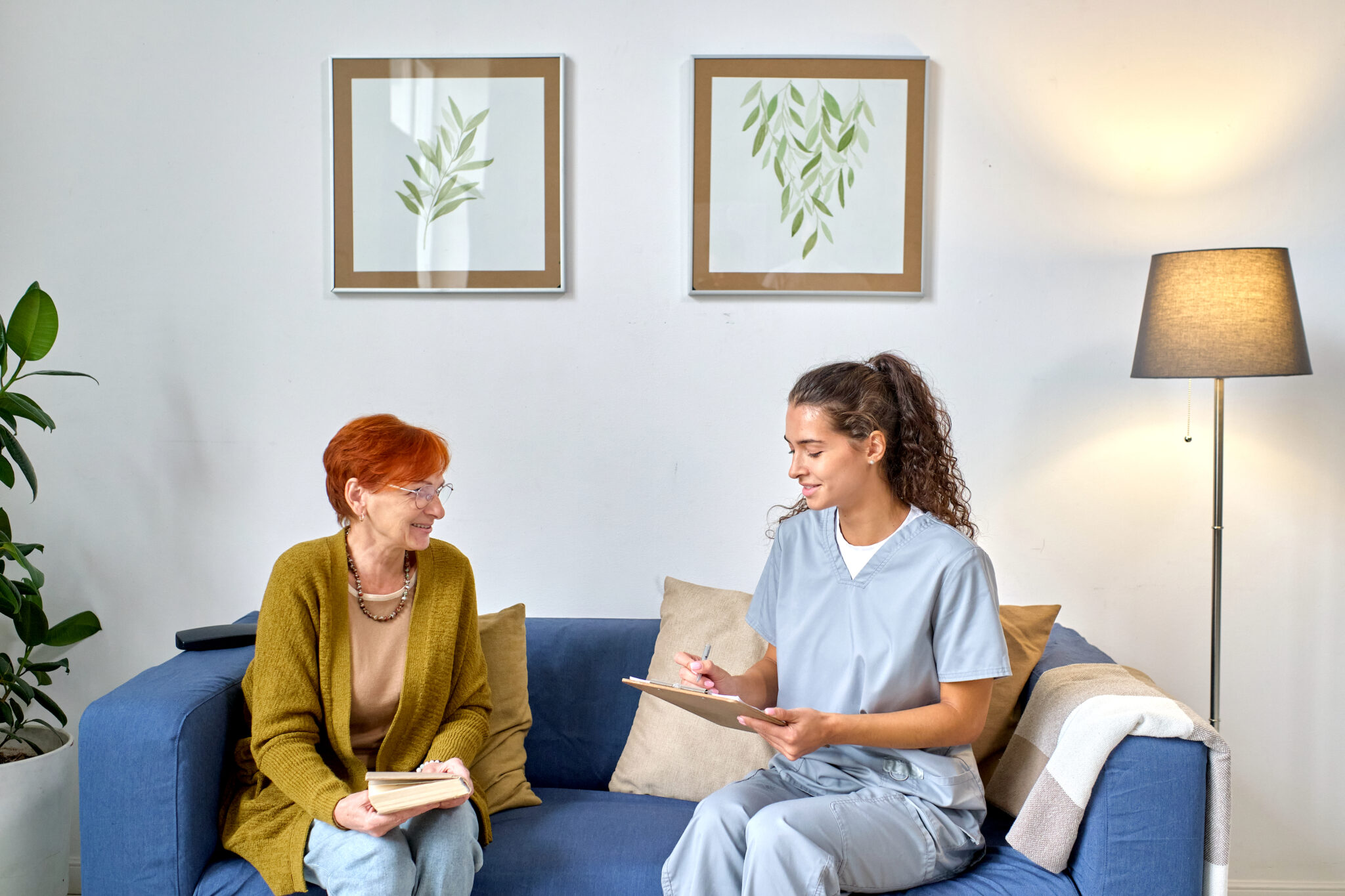 a home health care nurse and older adult female patient sitting together comfortably on a living room sofa and going over a list on a clipboard, possibly of supplies