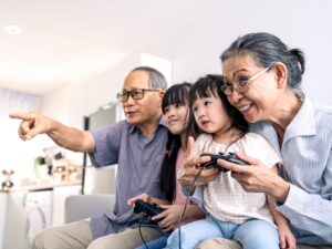 two small children with video game controllers sit on the laps of their grandparents aging in place in their apartment enjoying time playing together in the living room